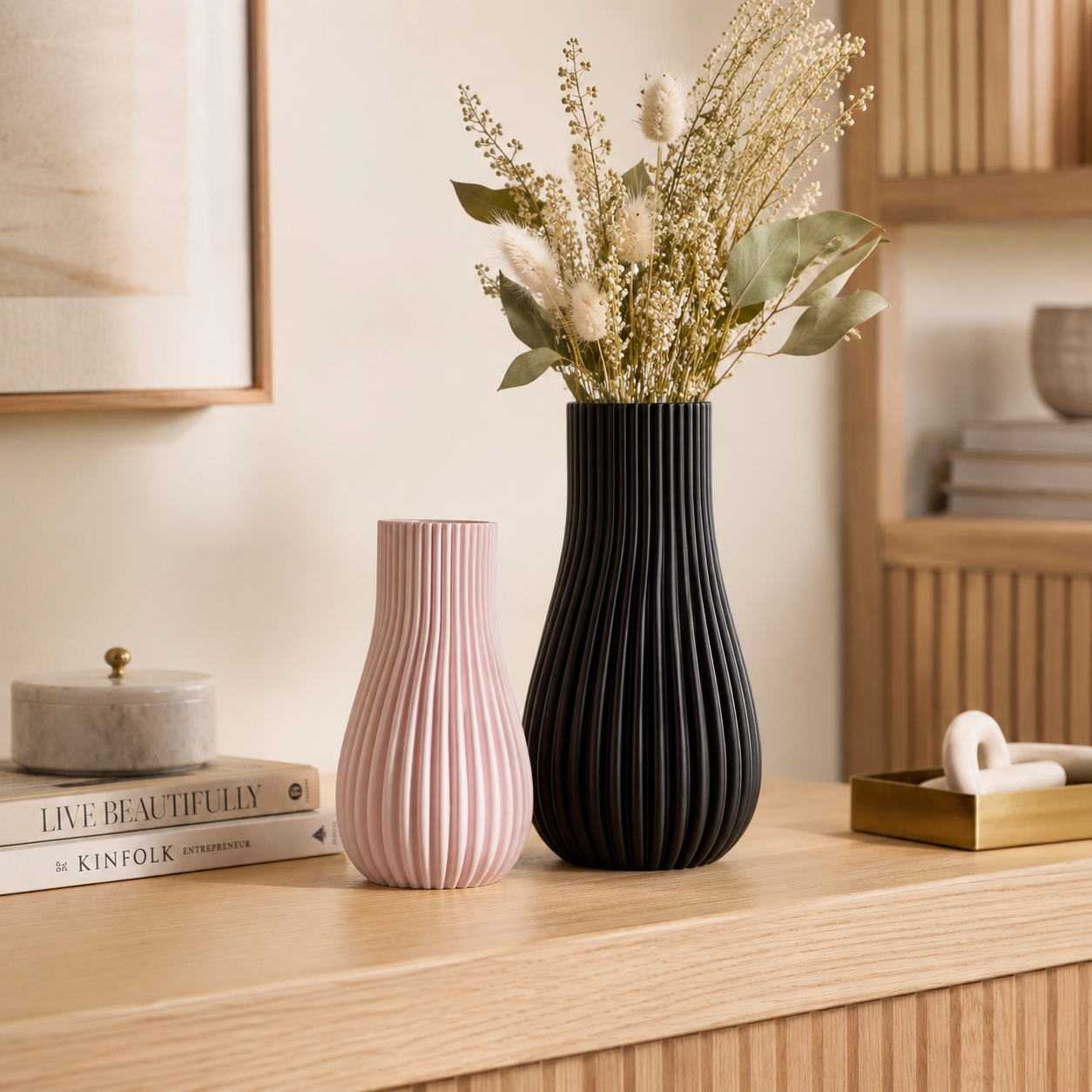 Two ribbed ceramic vases on a light wood console table, one in matte black and one in dusty pink, with dried flowers in the taller black vase. A framed print, books, and a small tray are visible in a softly styled interior setting.