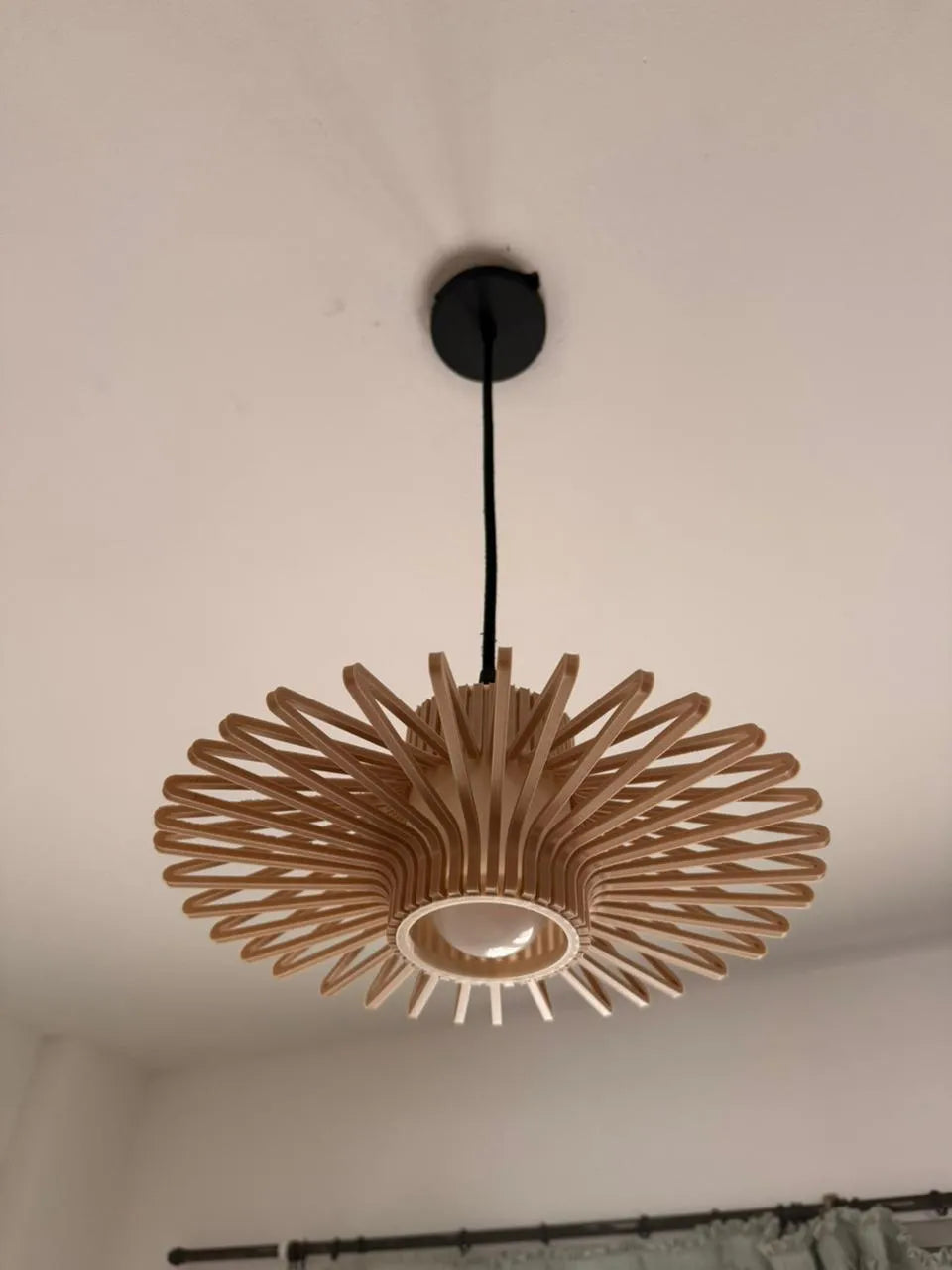 A modern radial pendant light made of natural wood slats, suspended from a black ceiling mount against a white ceiling.