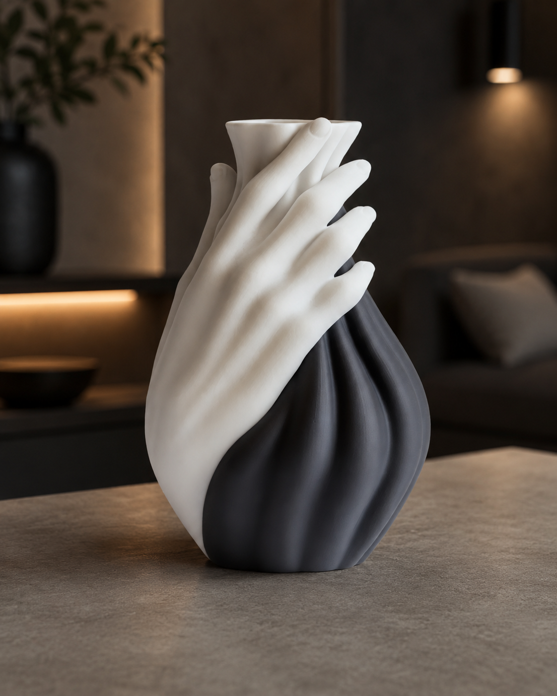 A sculptural vase shaped like two overlapping hands, with one half in matte white and the other in dark charcoal gray, displayed on a stone countertop in a modern, dimly lit interior setting.