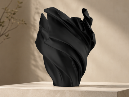 A sculptural black vase with flowing, draped folds, displayed on a light stone pedestal against a warm beige wall with soft shadows.
