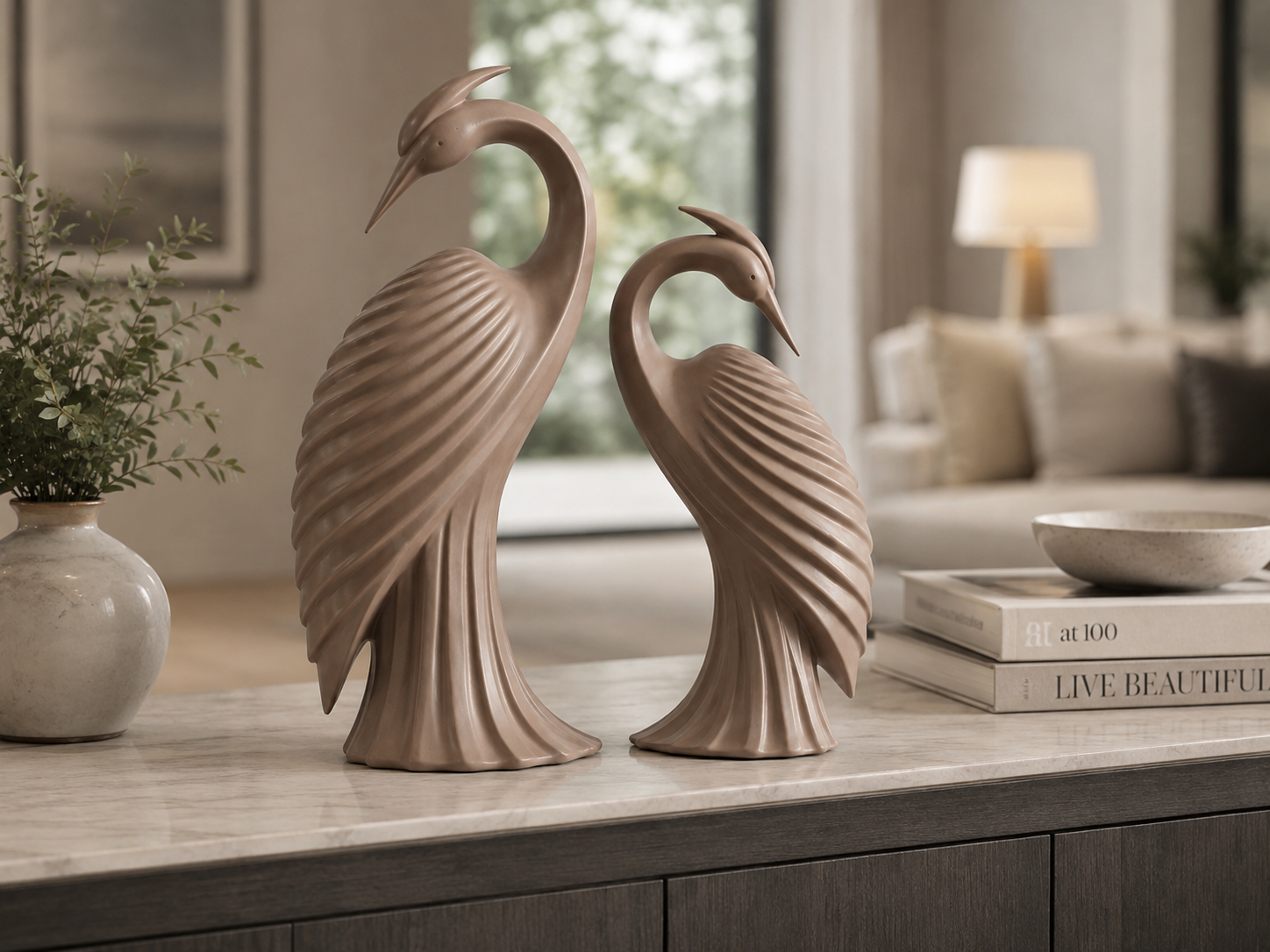Two decorative crane figurines in a warm taupe-beige tone, displayed side by side on a light marble console table in a softly blurred living room setting.