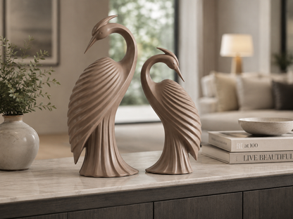 Two decorative crane figurines in a warm taupe-beige tone, displayed side by side on a light marble console table in a softly blurred living room setting.