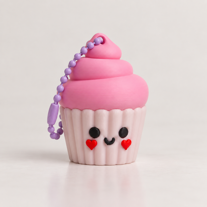 A cute cupcake-shaped keychain or accessory with pink frosting, a light pink wrapper, a smiling face, and a purple bead chain on a clean white background.
