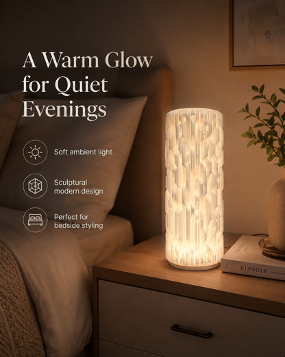 A cylindrical table lamp with a textured, ribbed white body glowing with a warm light, styled on a wooden nightstand next to a bed in a cozy bedroom setting.