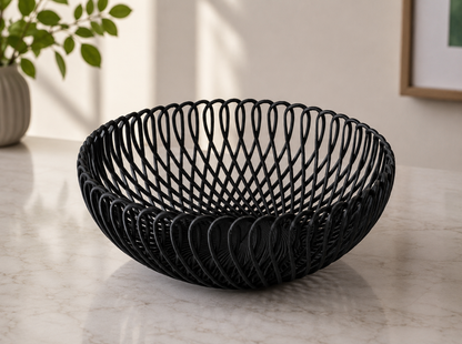 A black woven wire bowl with an open, spiral lattice design, displayed on a light marble countertop in a modern kitchen setting.