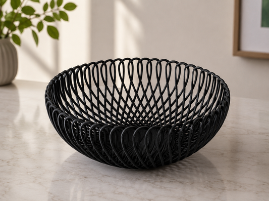 A black woven wire bowl with an open, spiral lattice design, displayed on a light marble countertop in a modern kitchen setting.