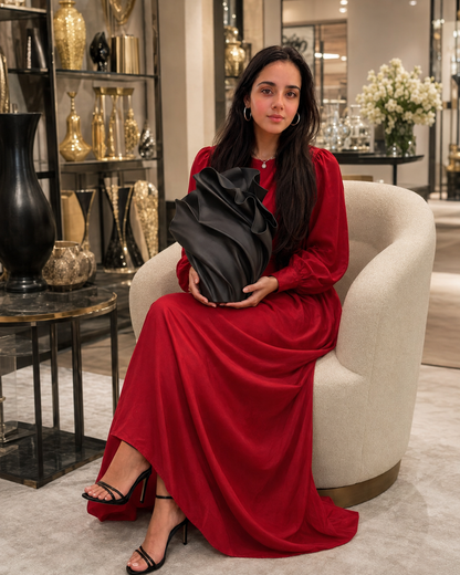 A woman with long dark hair sits in a cream-colored armchair inside a luxury boutique, wearing a long-sleeved red maxi dress and holding a black ruffled vase