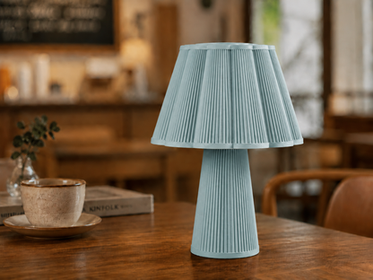 A light blue pleated table lamp with a flared shade and ribbed base, displayed on a wooden table in a warm, softly blurred indoor setting.