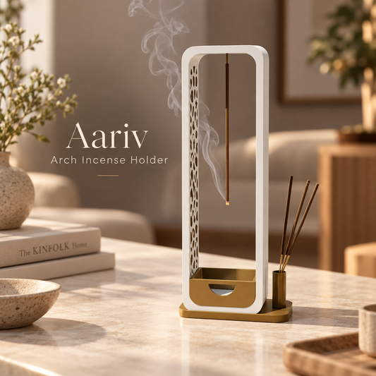 Aariv arch incense holder with a white rectangular frame and gold-toned base, shown on a marble table in a softly lit living room. Smoke rises from the hanging incense rod, and a small incense stick holder sits beside it.