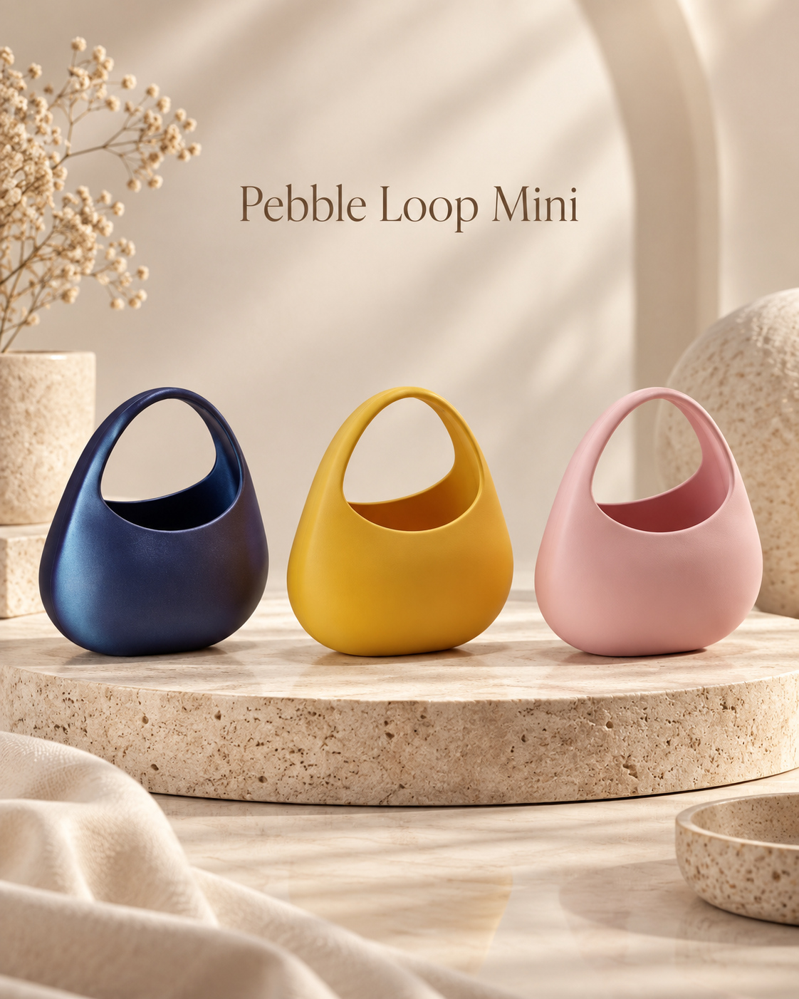 Three small Pebble Loop Mini handbags in navy blue, mustard yellow, and dusty pink, displayed on a round stone pedestal with neutral decor in the background.