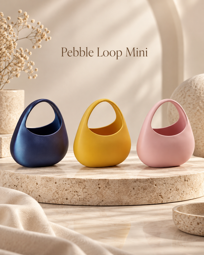 Three small Pebble Loop Mini handbags in navy blue, mustard yellow, and dusty pink, displayed on a round stone pedestal with neutral decor in the background.