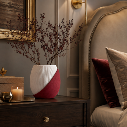 A modern ceramic vase with a diagonal split design of white ribbed texture and deep red suede-like fabric, holding dried branches, styled on a dark wood nightstand next to a bed in a warm, elegant bedroom.