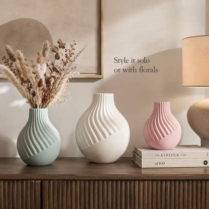 Three ribbed ceramic vases in sage green, off-white, and blush pink are styled on a wooden cabinet against a beige wall. The vases are decorated with dried grasses and books, with a lamp and framed art in the background.