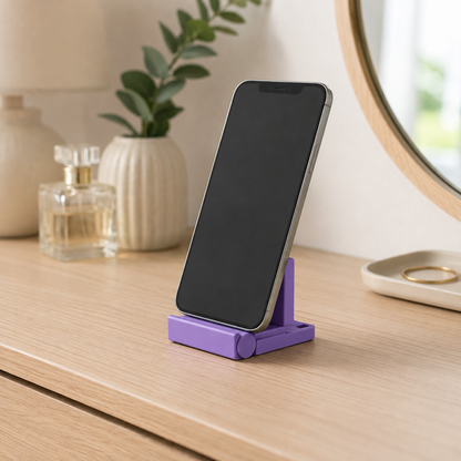 A purple folding smartphone stand holding a black smartphone, set on a light wood tabletop in a styled indoor scene with a vase, perfume bottle, and mirror in the background.