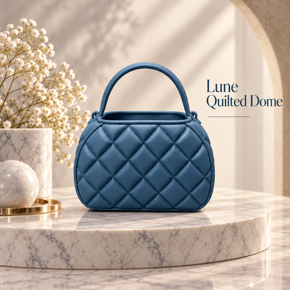 A blue quilted dome-shaped handbag with a top handle, displayed on a round marble pedestal. The background features a warm beige archway, soft shadows, and a vase of small white flowers.