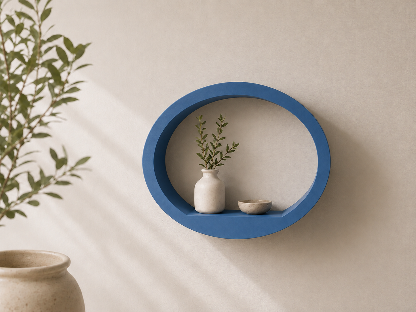 A blue oval floating wall shelf mounted on a beige wall, styled with a small white ceramic vase and a dried green plant, with a beige ceramic pot partially visible in the foreground.