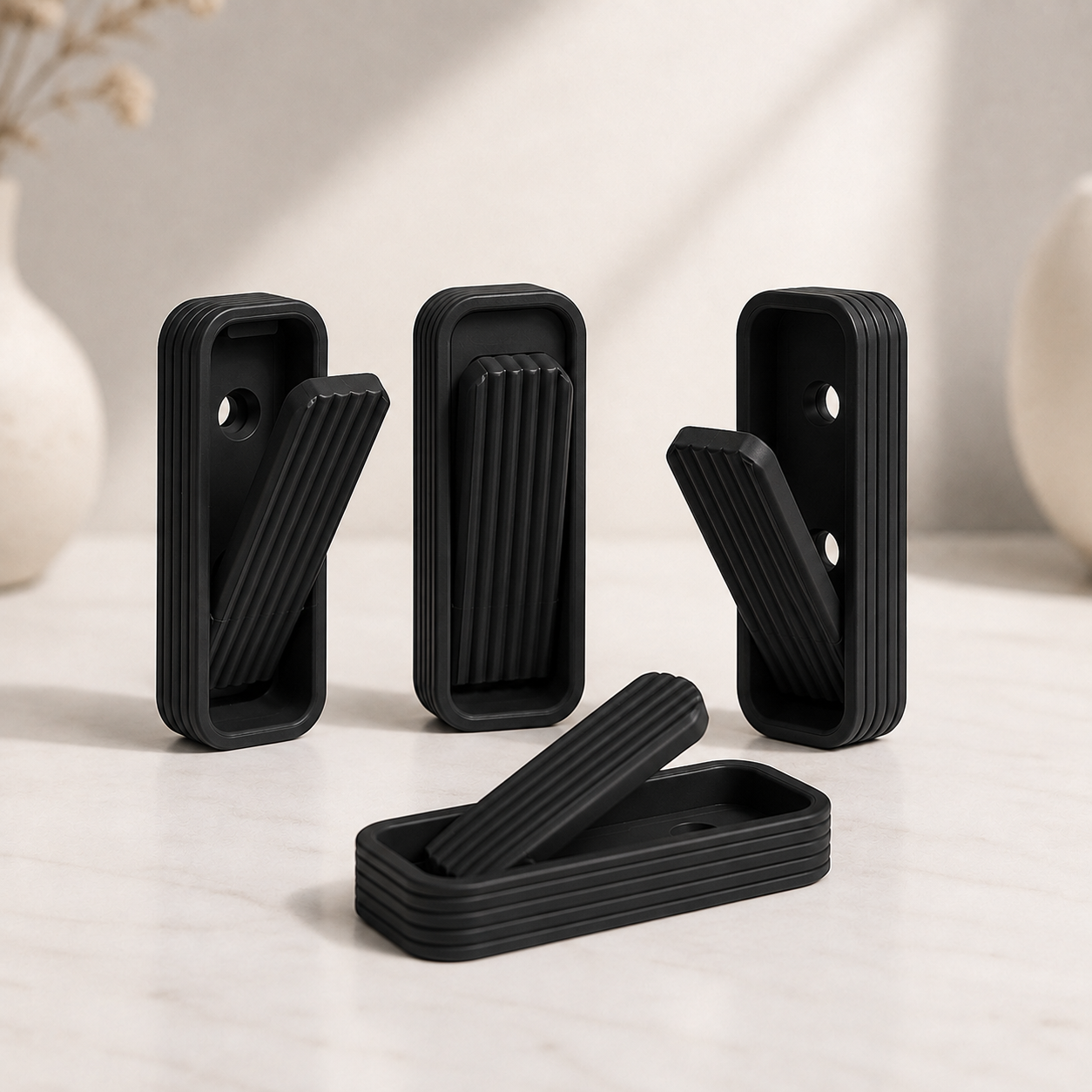 Four black rectangular wall hooks with ribbed handles, shown in both open and closed positions on a light marble surface with soft-focus decorative vases in the background.