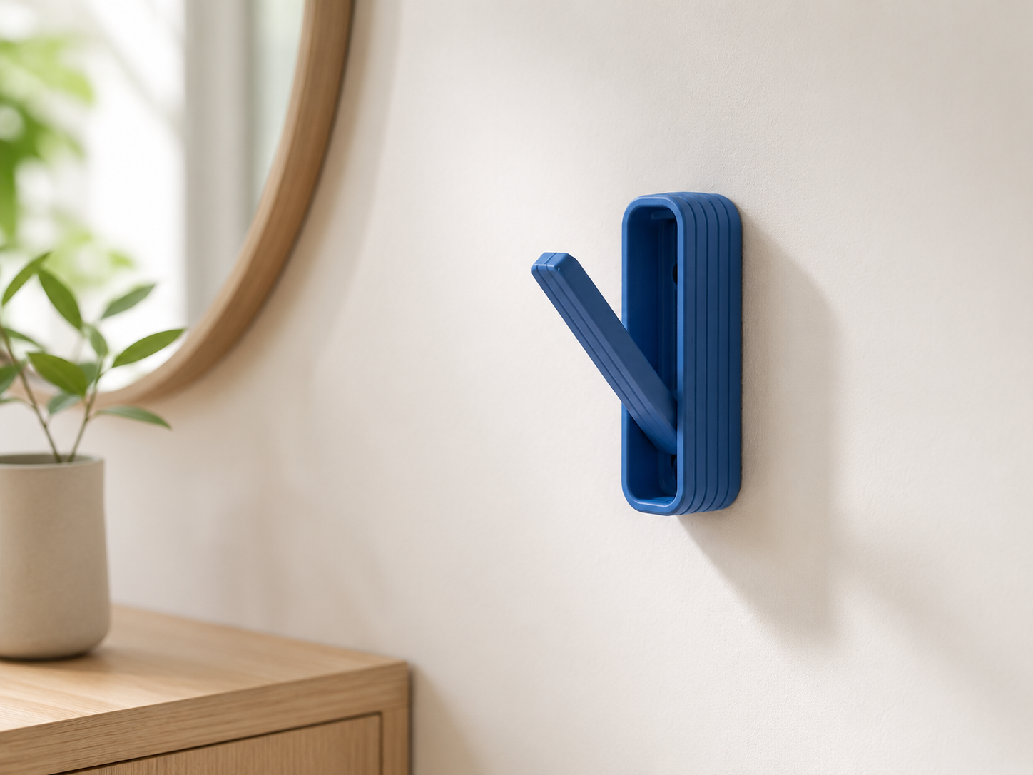 A blue silicone wall hook with a textured, ribbed design and a hinged arm, mounted on a warm off-white wall in a modern interior setting.
