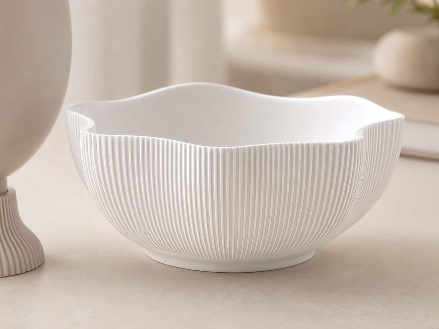 A white ceramic bowl with a wavy rim and vertical fluted texture, shown on a light beige tabletop with a softly blurred background.