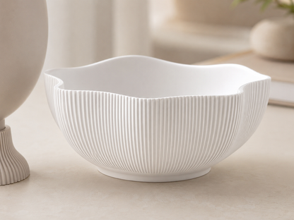 A white ceramic bowl with a wavy rim and vertical fluted texture, shown on a light beige tabletop with a softly blurred background.