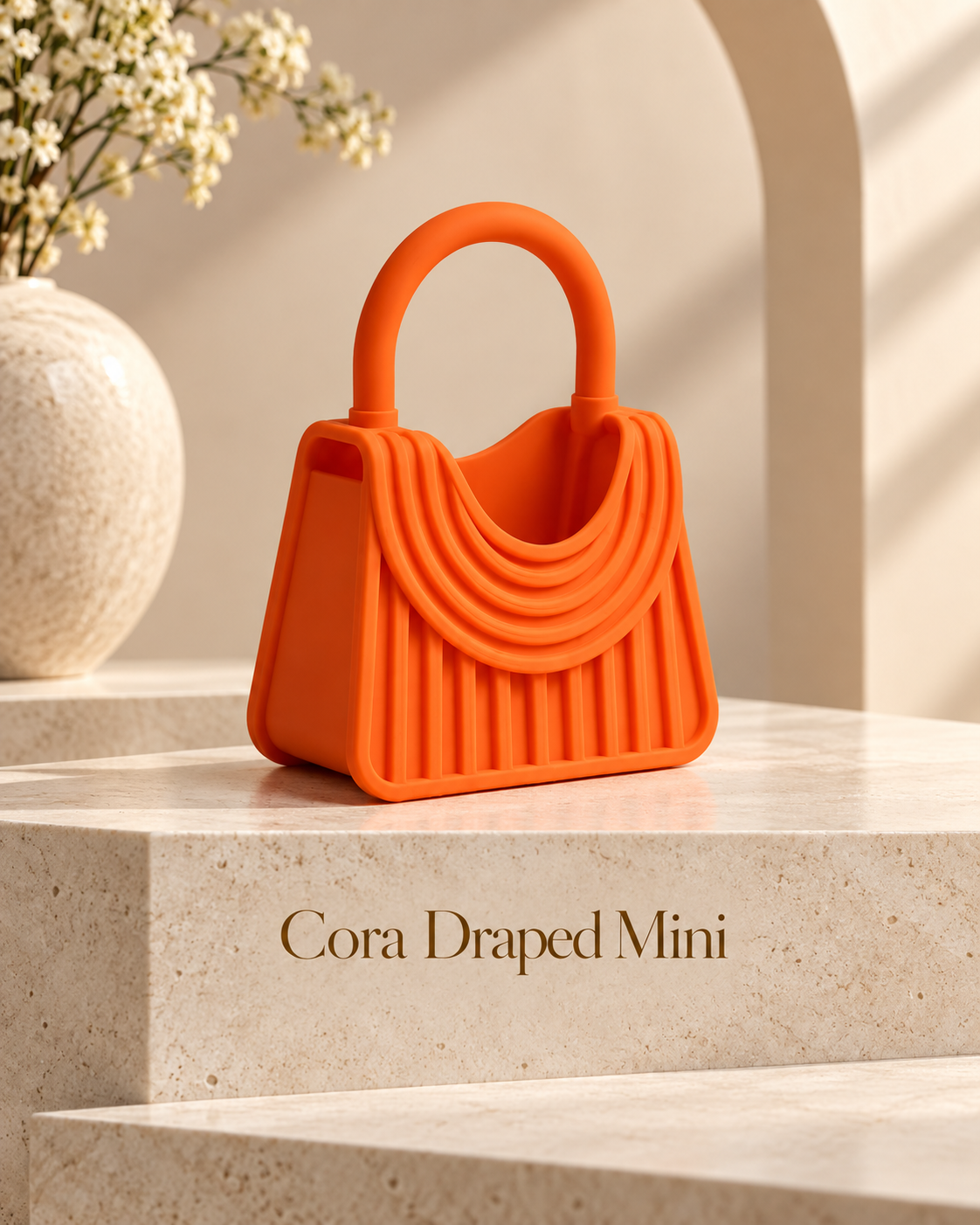 An orange silicone handbag-shaped object labeled 'Cora Draped Mini' sits on a beige stone pedestal in a styled interior scene with a vase of small white flowers and warm directional light.