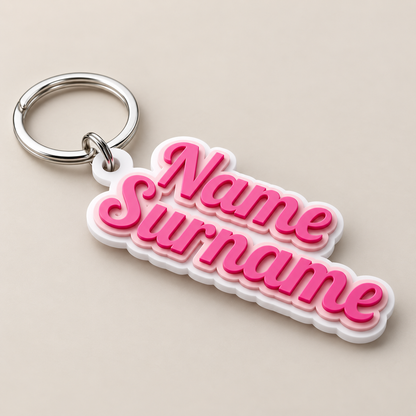 A customizable keychain featuring the words 'Name Surname' in 3D pink bubble letters with a white outline, attached to a silver metal split ring, set against a solid beige background.