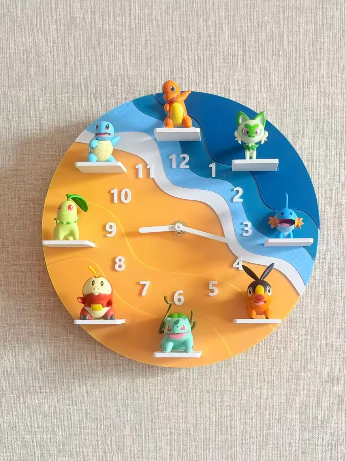 A round wall clock featuring colorful Pokémon characters mounted on white platforms at each hour position, with a blue and orange wavy face design on a beige textured wall.