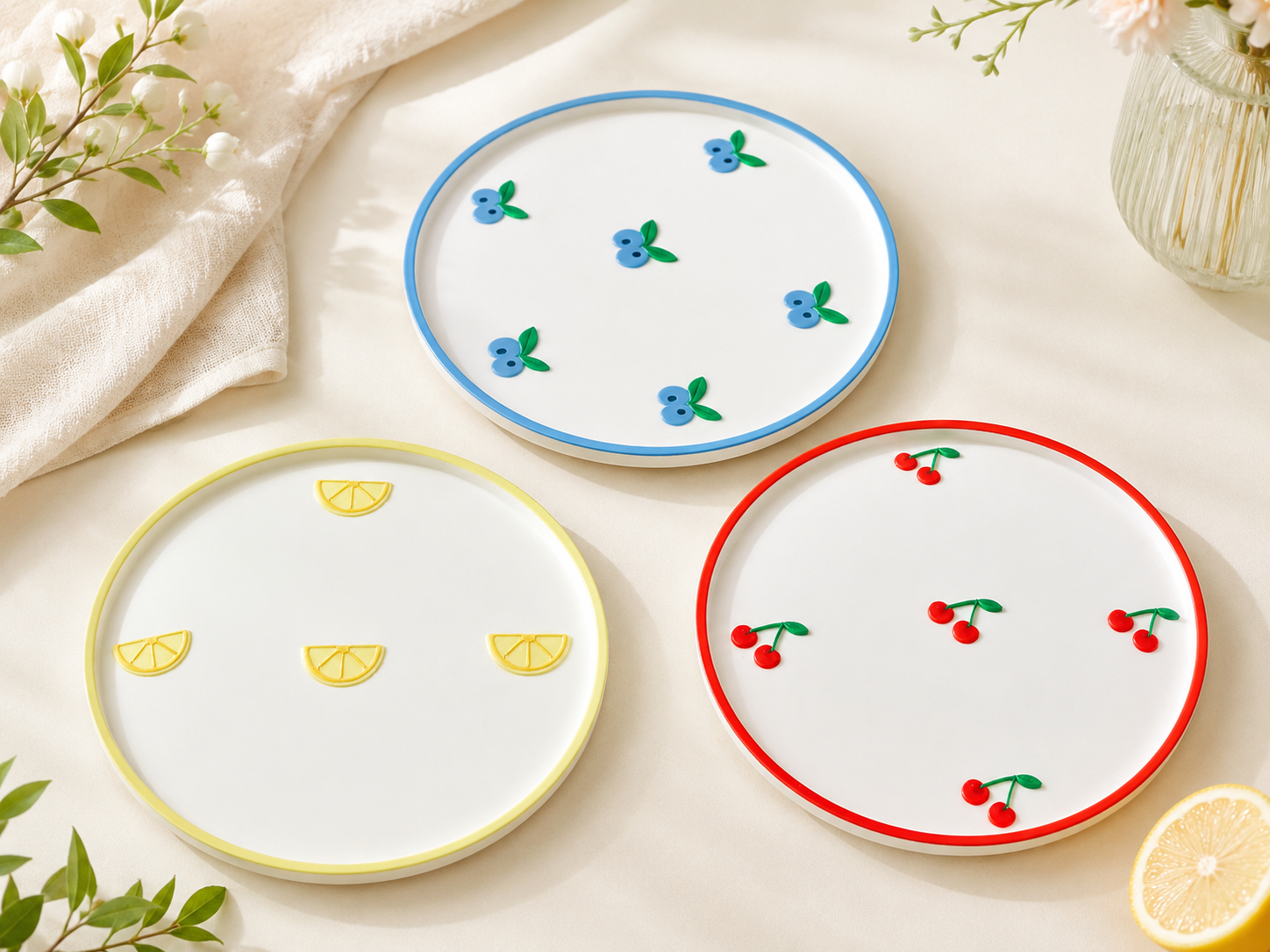 Three round white ceramic plates with colorful borders and fruit motifs, arranged on a light cream surface with decorative leaves, fabric, and a glass vase.