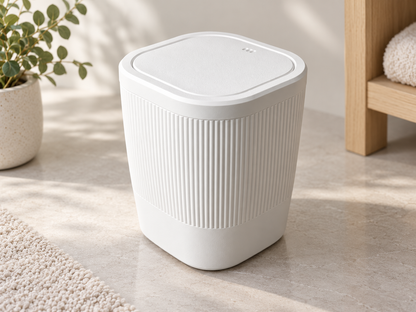 A white modern cylindrical trash can with a ribbed front and a square-shaped lid, shown in a bright, sunlit room setting on a light stone floor.