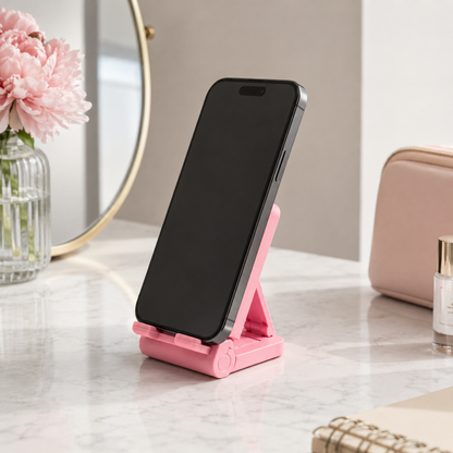 A pink folding smartphone stand holding a black smartphone, set on a light marble desk with a pink peony in a glass vase, a round mirror, and a blush pouch in the background.