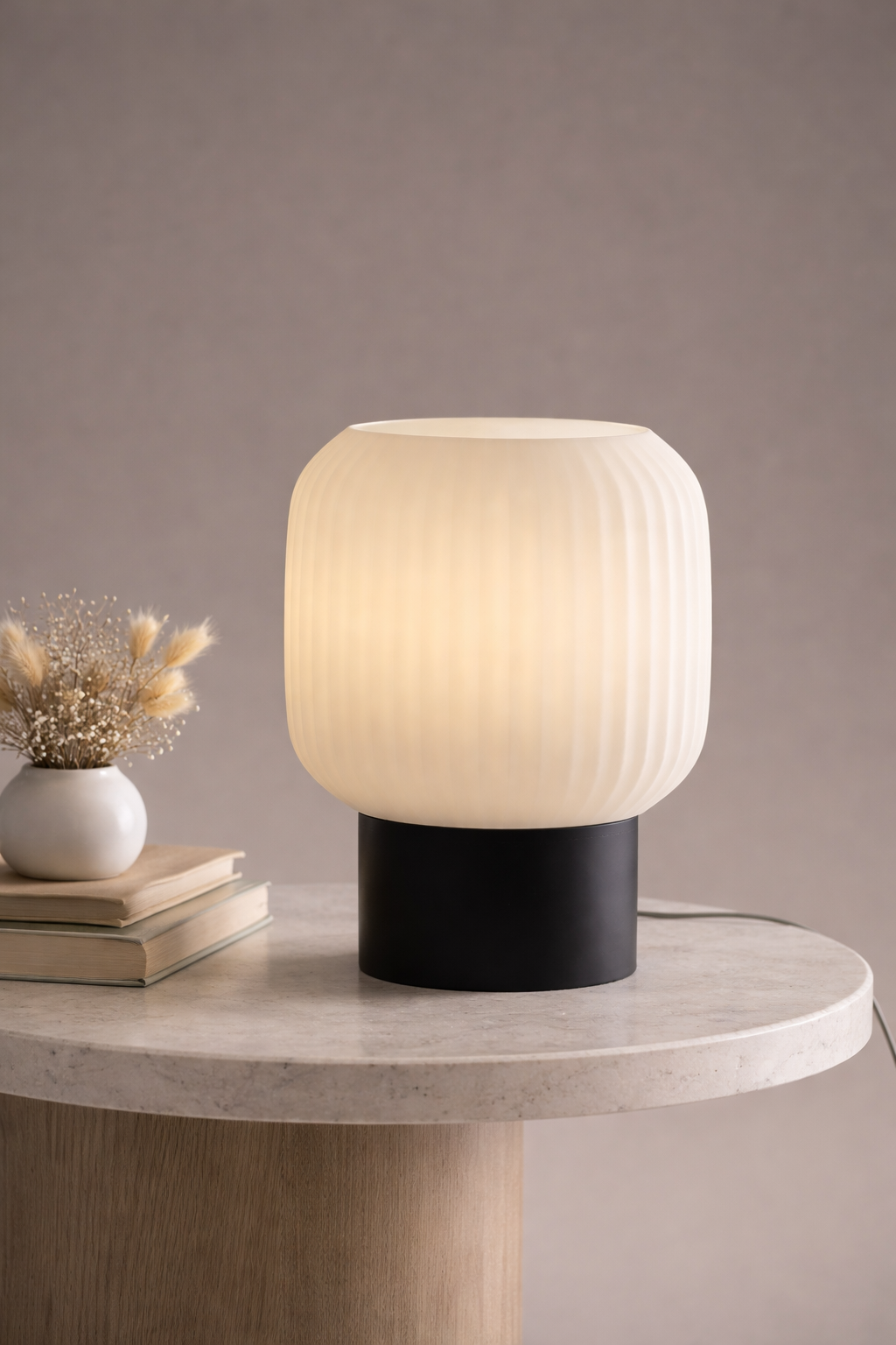 Table lamp with ribbed white shade and black base on a marble surface with books and a vase.