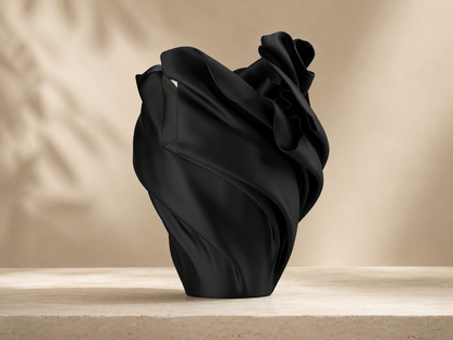 A sculptural black vase made from draped, flowing fabric, displayed on a light beige stone surface against a warm beige background with soft leaf shadows.