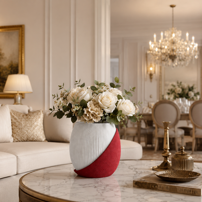 A modern ceramic vase with a diagonal split design in white and red, filled with white roses and green foliage, placed on a marble-topped table in a luxurious, softly lit living room.