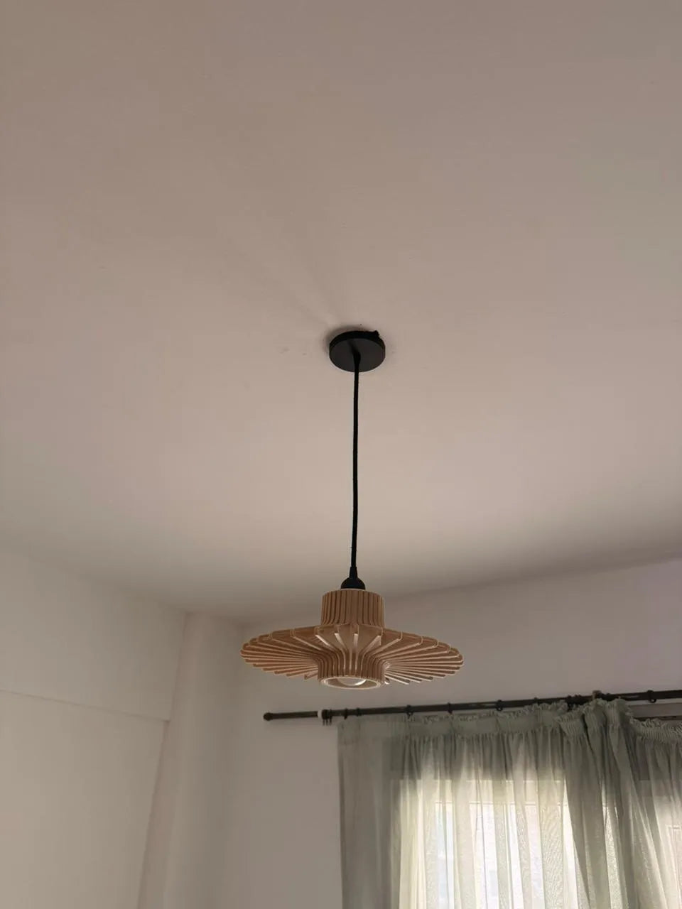 A modern pendant light with a natural wood slatted shade and a black cord, hanging from a black ceiling mount on a white ceiling.