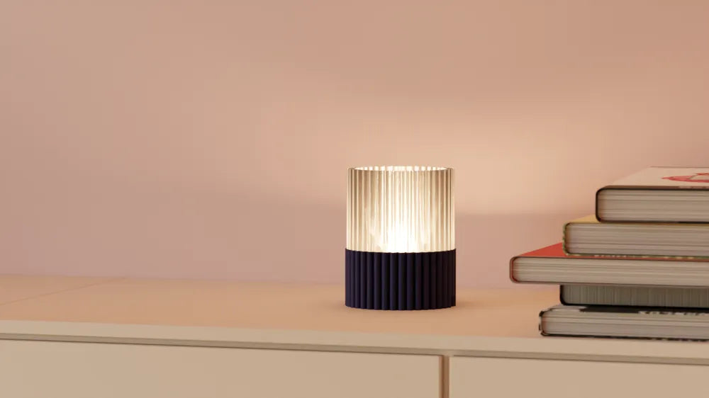 A modern pleated table lamp with a ribbed dark navy base and a glowing pleated shade, styled on a light beige shelf next to a stack of books against a soft pink wall.