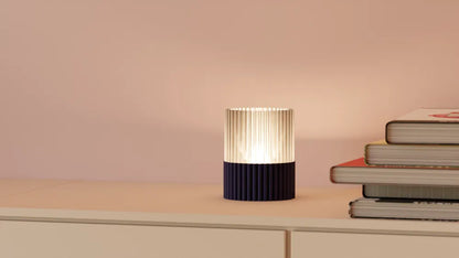 A modern pleated table lamp with a ribbed dark navy base and a glowing pleated shade, styled on a light beige shelf next to a stack of books against a soft pink wall.