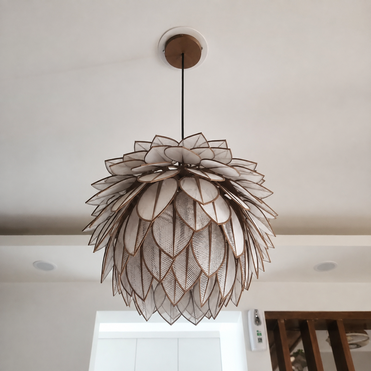 A modern flower-shaped pendant light with layered beige and white mesh petals outlined in dark brown, hanging from a black cord on a white ceiling.
