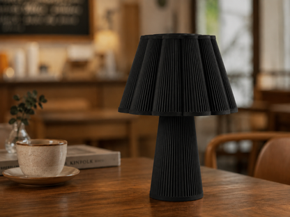 A black pleated table lamp with a flared shade and ribbed base, displayed on a wooden table in a warm, softly blurred indoor setting.