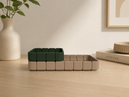 A modern modular desk organizer made of square plastic bins, shown in two colors: dark forest green and light beige. The organizer is styled on a light wood desk with a ceramic vase, a framed print, and a book in the background.
