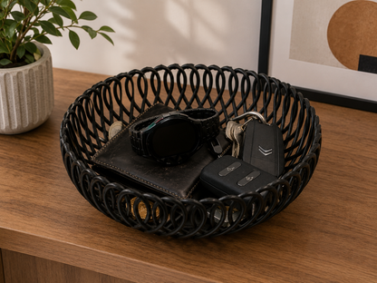 A round black metal woven basket tray sits on a wooden console table, styled with a wallet, smartwatch, keys, and a car key fob. A potted plant and framed wall art are visible in the background.