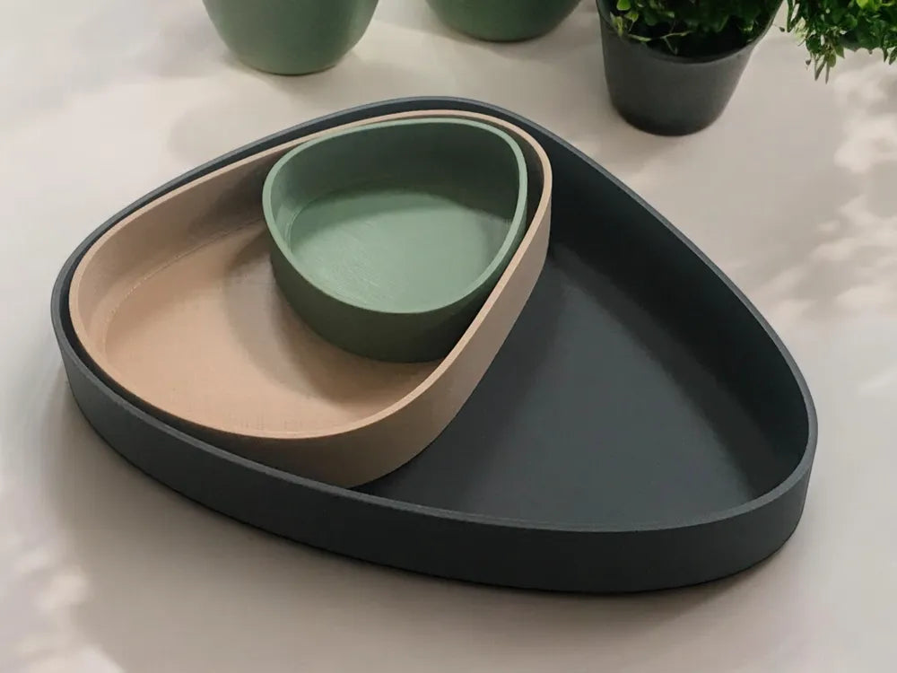 Three nested modern trays in matte dark gray, beige, and sage green, arranged on a light surface with potted plants blurred in the background.