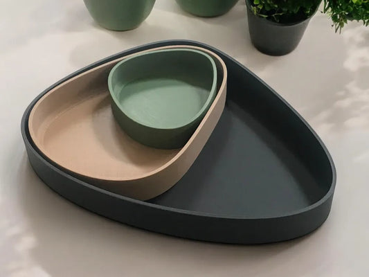 Three nested modern trays in matte dark gray, beige, and sage green, arranged on a light surface with potted plants blurred in the background.