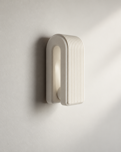 A modern wall-mounted light fixture with a ribbed, arched rectangular design in a warm off-white finish, shown mounted on a textured wall with soft shadows.