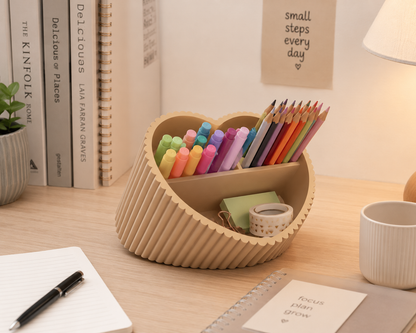 A heart-shaped wooden desk organizer with a slatted ribbed exterior and two compartments, filled with colored pencils, highlighters, and small stationery items, placed on a light wood desk with books, a mug, and a lamp in the background.