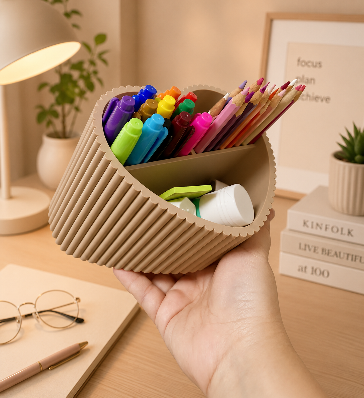 A hand holding a ribbed, heart-shaped beige desk organizer filled with colorful pens, pencils, and sticky notes on a warm, styled desk background.