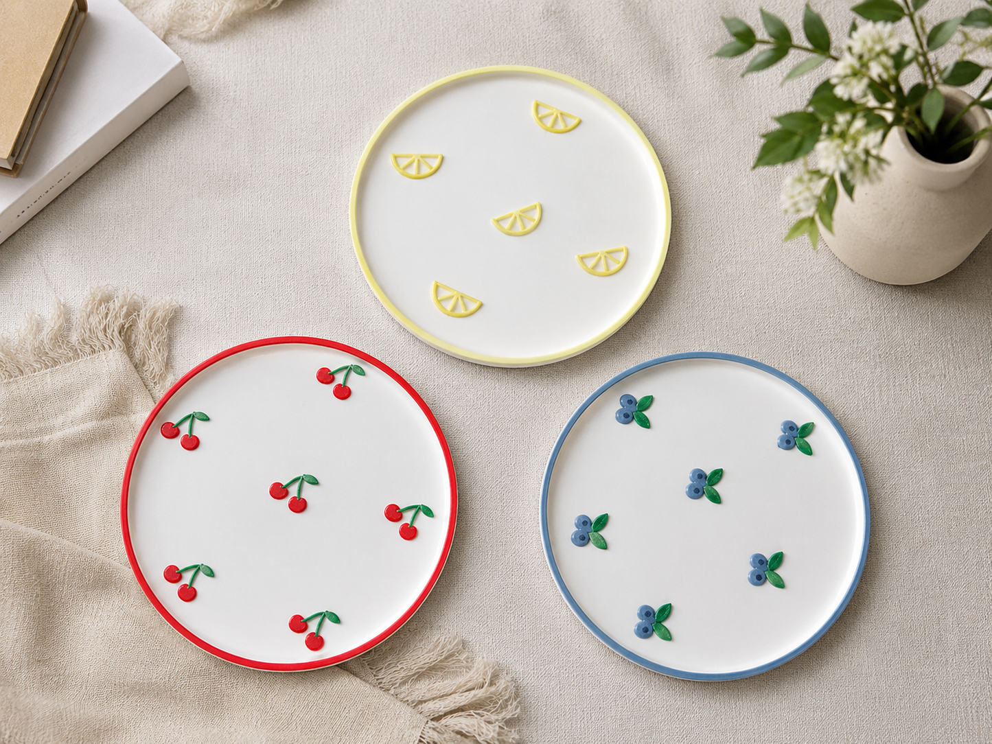 Three round white trays with lemon, cherry and blueberry fruit motifs arranged on a beige fabric surface