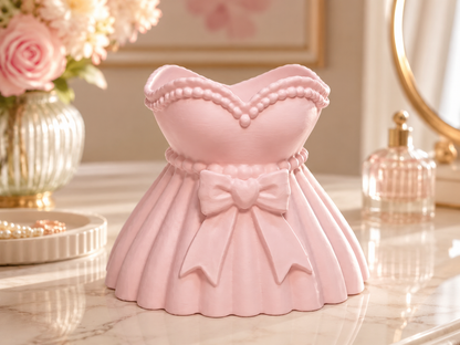 A pink dress-shaped decorative container or organizer displayed on a marble surface in a softly styled room setting with flowers, a mirror, and a perfume bottle.