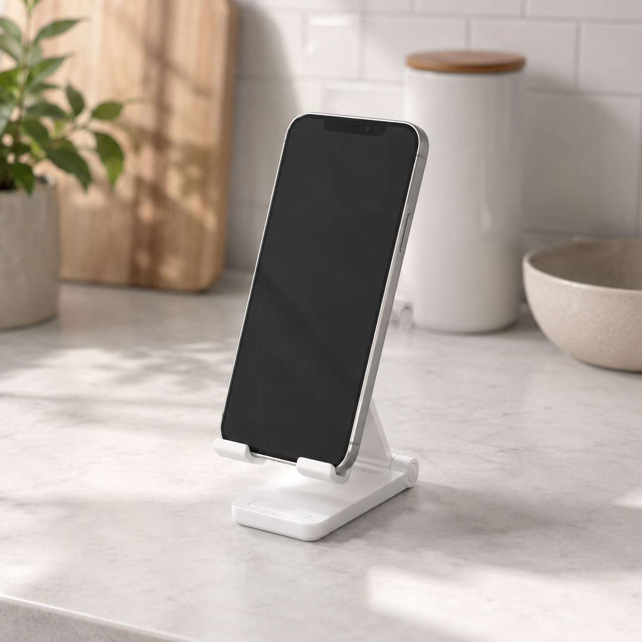 A white minimalist smartphone stand holding a black smartphone, set on a light marble countertop in a bright kitchen environment.