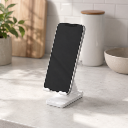 A white minimalist smartphone stand holding a black smartphone, set on a light marble countertop in a bright kitchen environment.