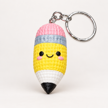 A cute, crocheted pencil-shaped keychain with a smiling face, featuring a pink top, yellow body, white lower section, black tip, and a silver metal key ring.